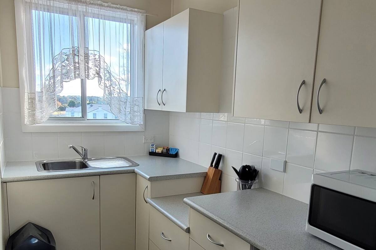 Cocina compacta con armarios blancos, microondas, lavamanos y ventana con cortinas traslúcidas.