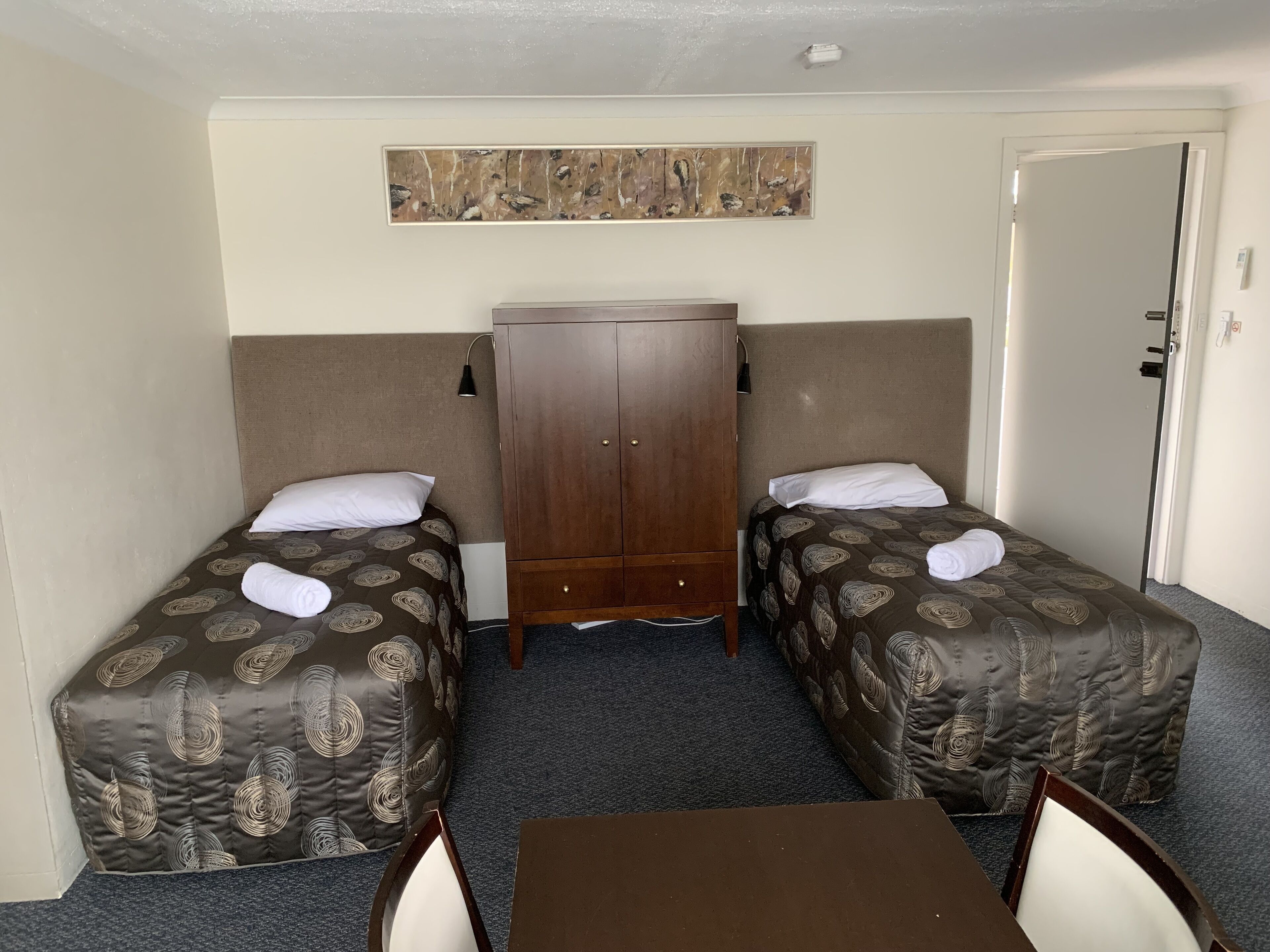 Una habitación de hotel con dos camas individuales, un armario de madera, una mesa y una obra de arte colgada en la pared.
