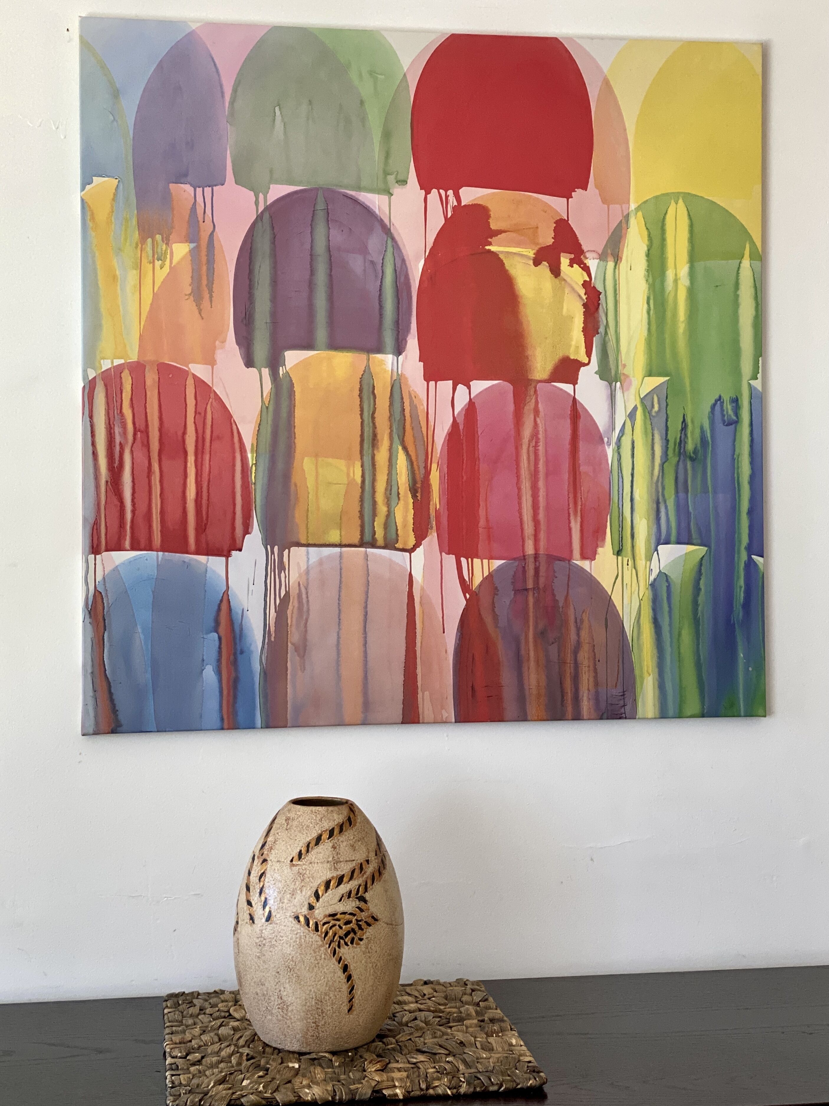 A colorful abstract painting on a wall with a textured vase on a woven placemat below.