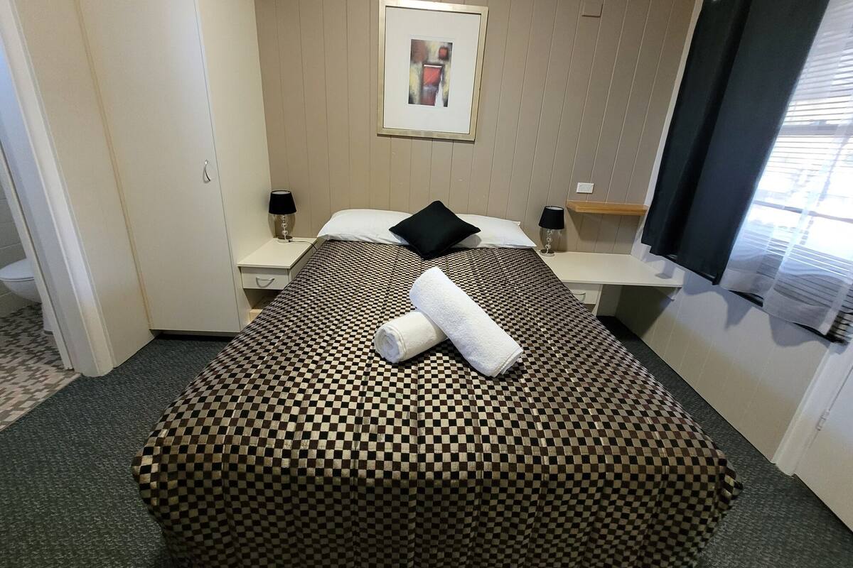 Habitación de hotel con colcha a cuadros, almohada, toalla enrollada, mesita de noche con lámpara y un cuadro en la pared.