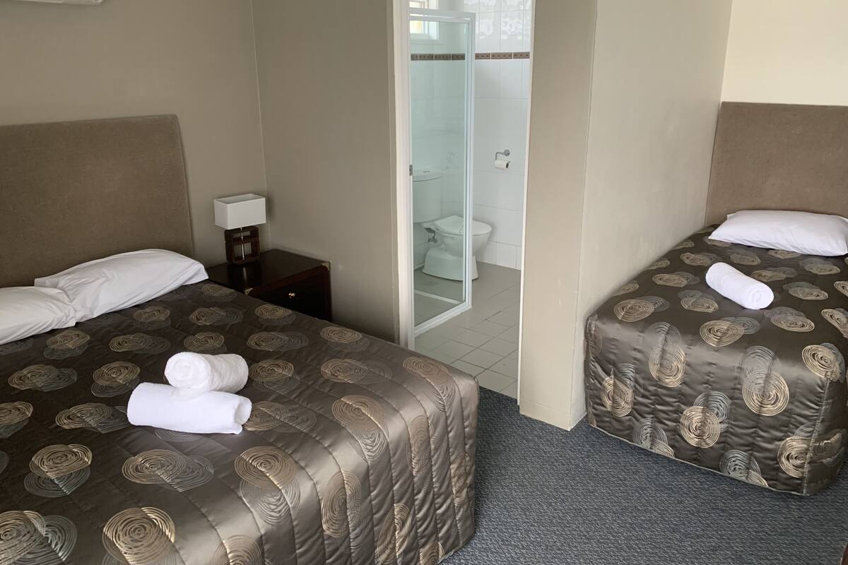 Habitación de hotel con dos camas, una mesita de noche, una lámpara y un baño visible a través de una puerta de cristal.
