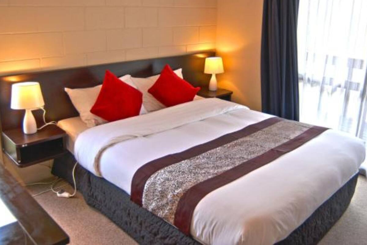 A hotel room with a large bed, two red pillows, a white comforter, and a dark headboard. There are two bedside lamps and a window with curtains.