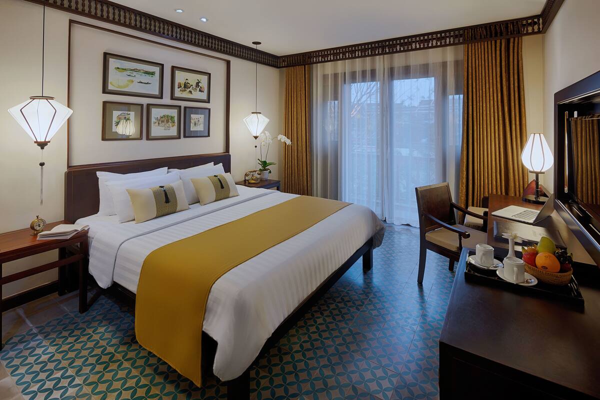 Hotelkamer met een groot bed, een bureau met een laptop, een stoel en een dienblad met kopjes en fruit.