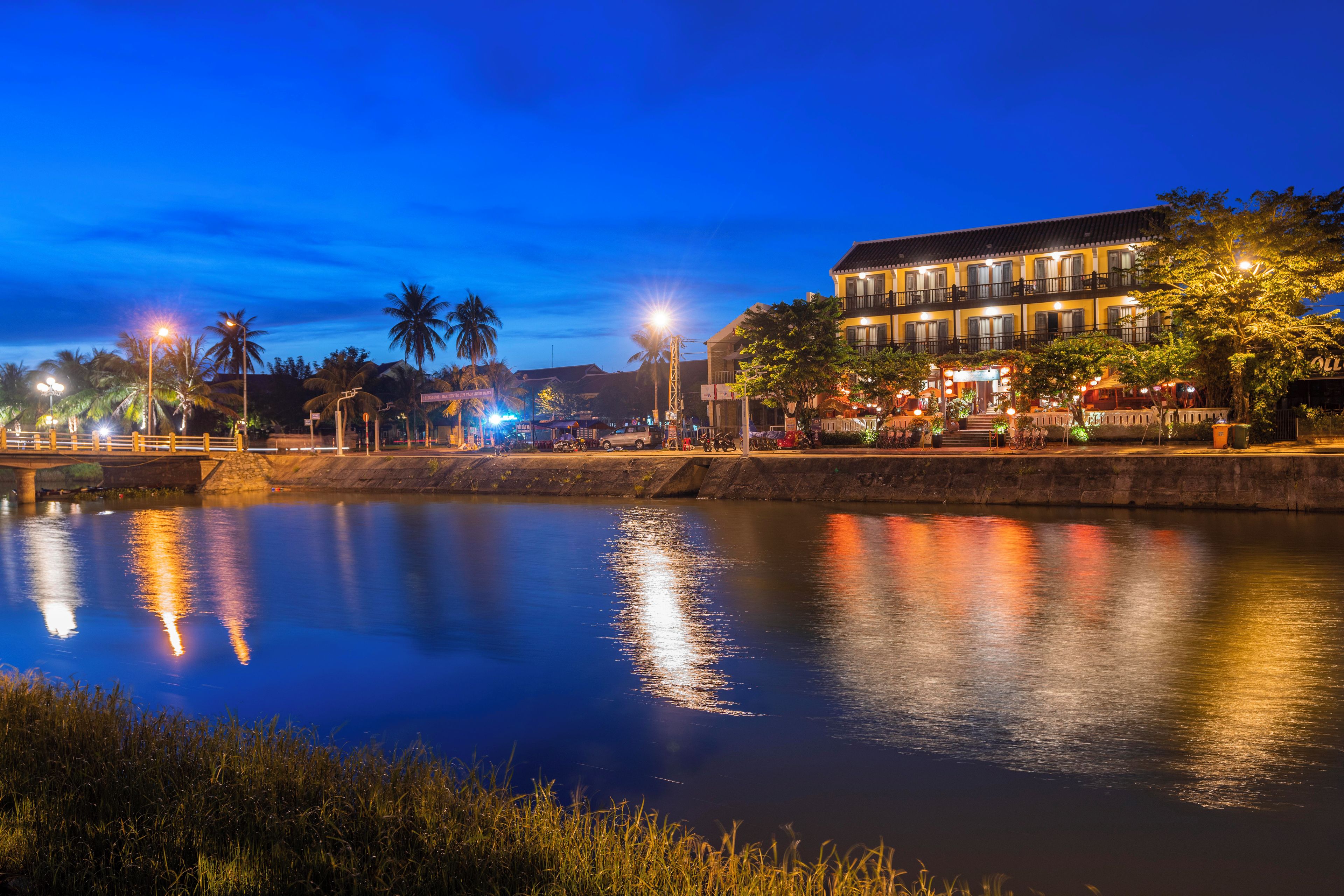 A waterfront hotel with a reflection of the building in the water, palm trees, and a bridge in the background.