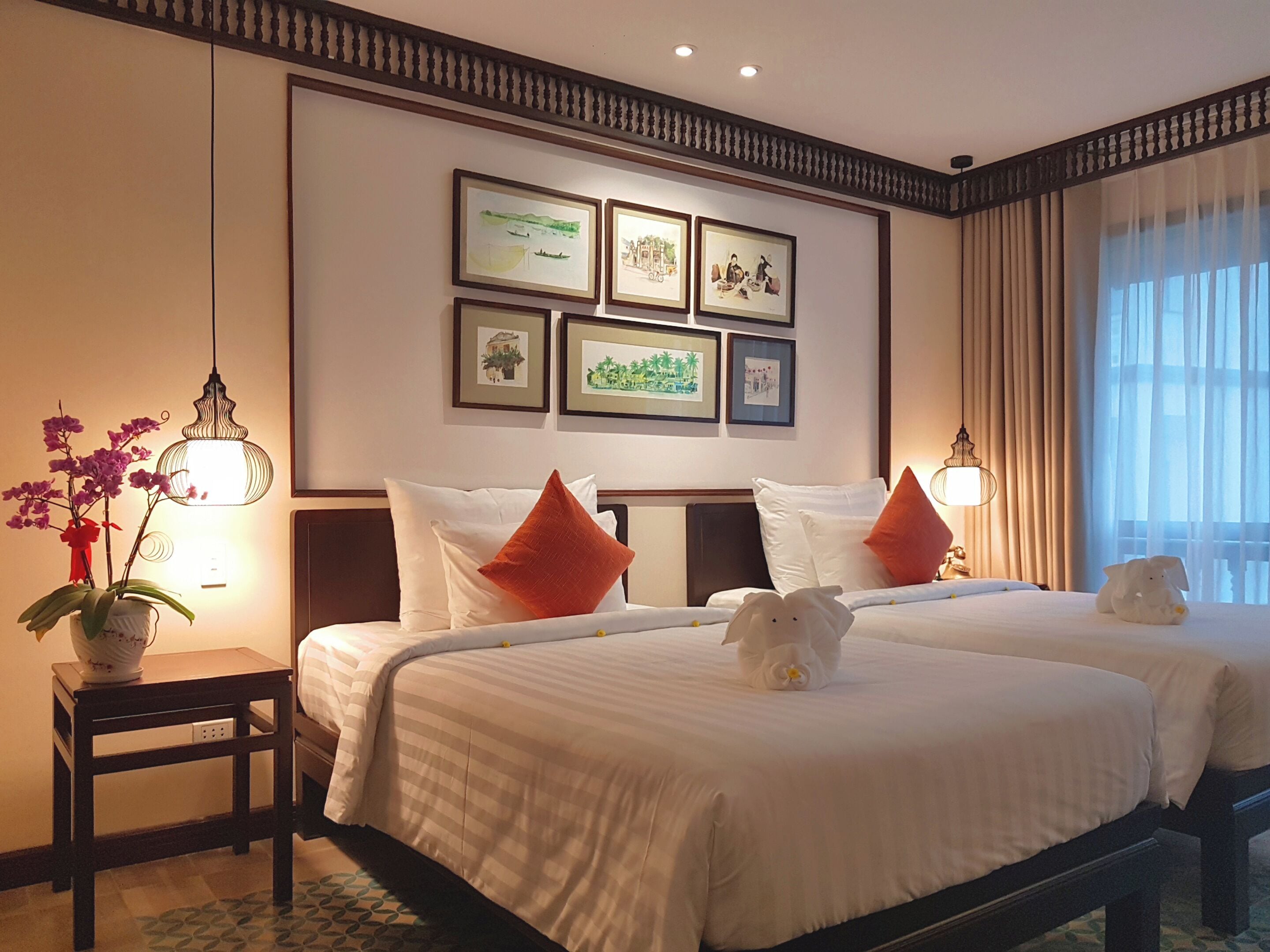 Habitación de hotel con dos camas, una mesita con un florero, cuadros en la pared y una ventana con cortinas.