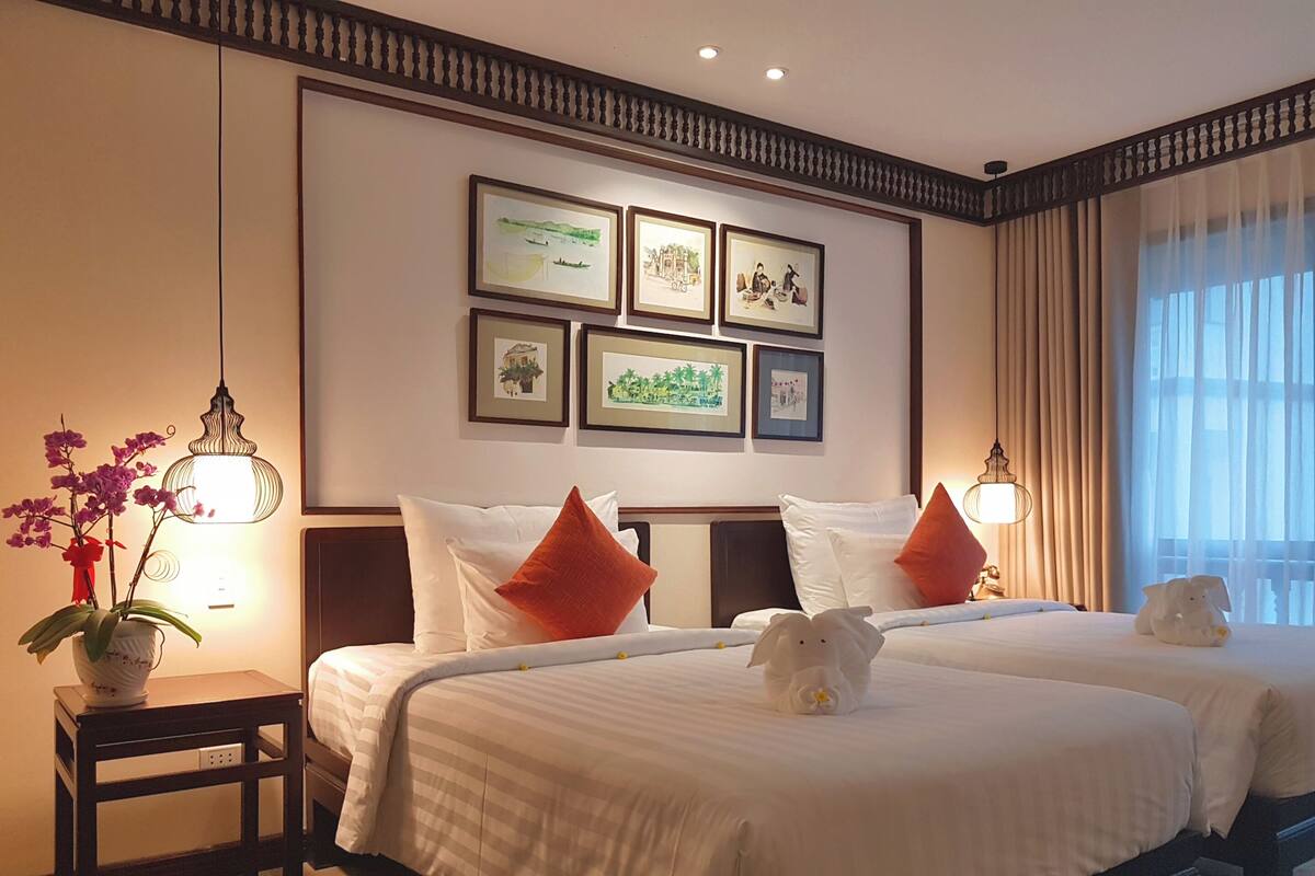 Een hotelkamer met twee bedden, een klein tafeltje met een bloemenvaas, prestaties aan de muur en een raam met gordijnen.