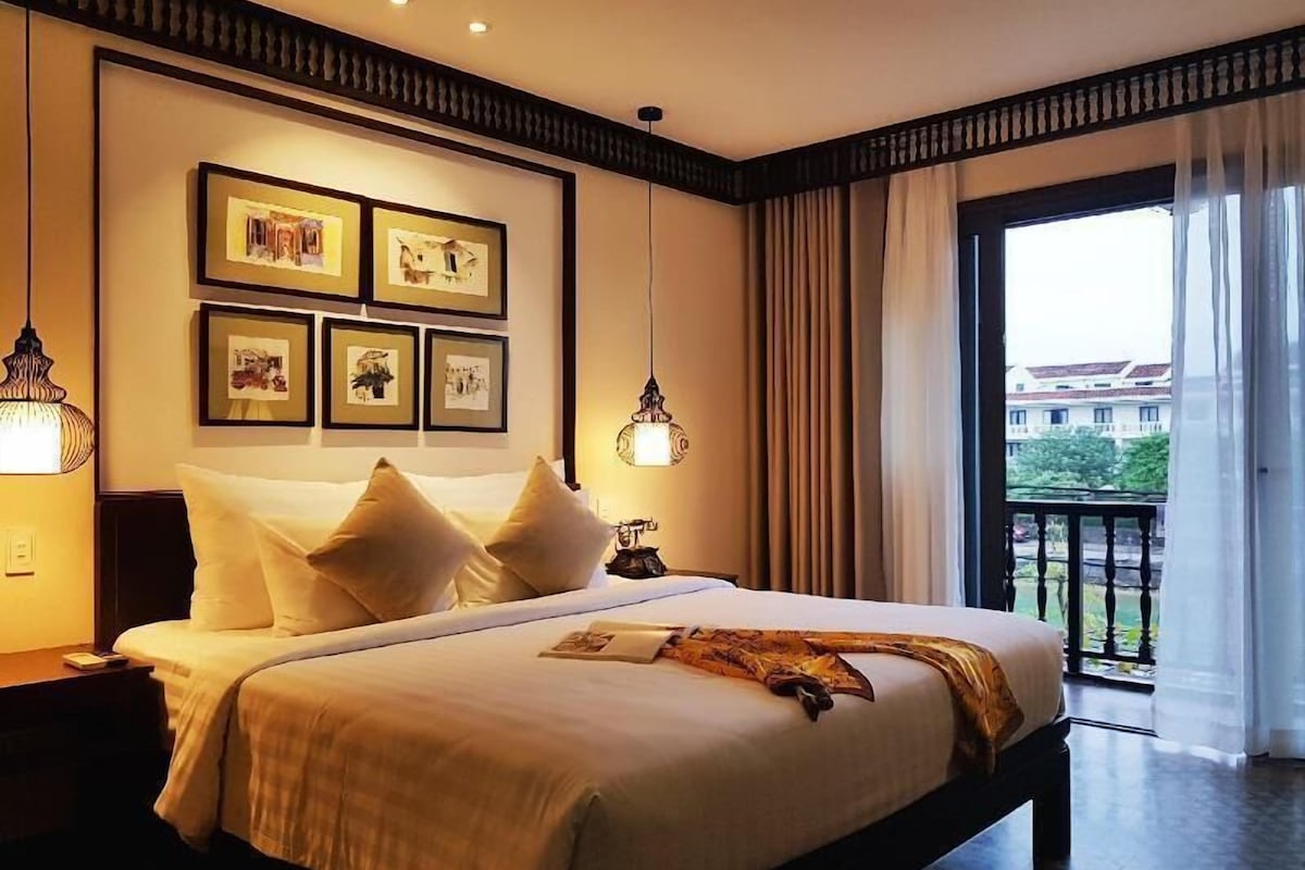 Ein Hotelzimmer mit einem großen Bett, gerahmten Bildern an der Wand, einem Balkon mit Vorhängen und Blick auf umliegende Gebäude.