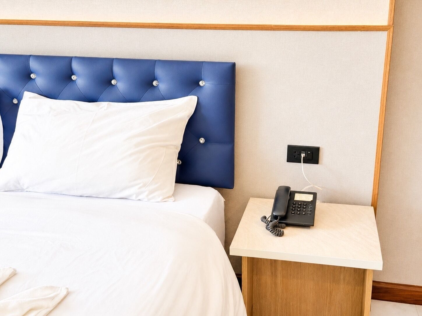 Une chambre d’hôtel avec une tête de lit bleue à motifs capitonnés, du linge de lit blanc, une table de chevet avec un téléphone noir et une table de nuit en bois.