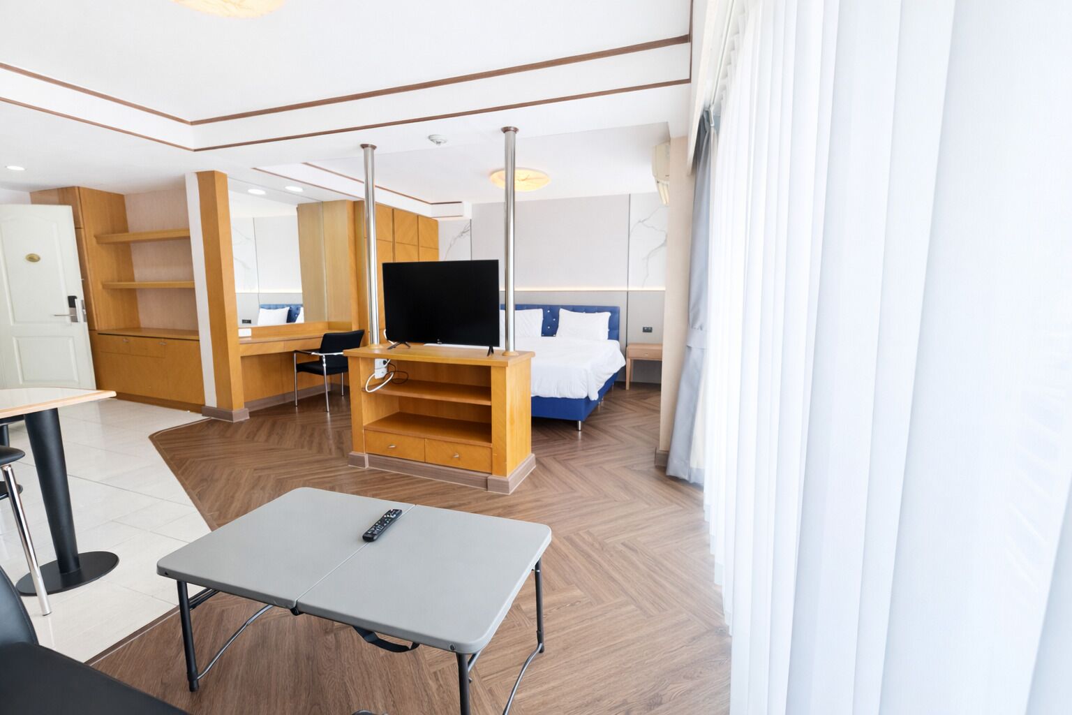 A hotel room with a bed, a TV on a wooden stand, a small table, a chair, and a desk with a remote control.