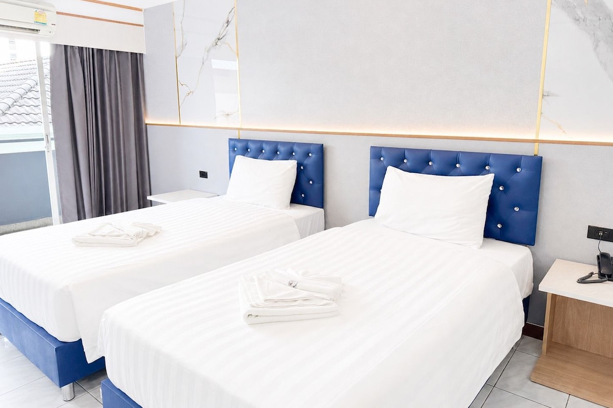 Habitación de hotel con dos camas, una ventana con cortinas, un equipo de aire acondicionado y una mesita de noche con lámpara.