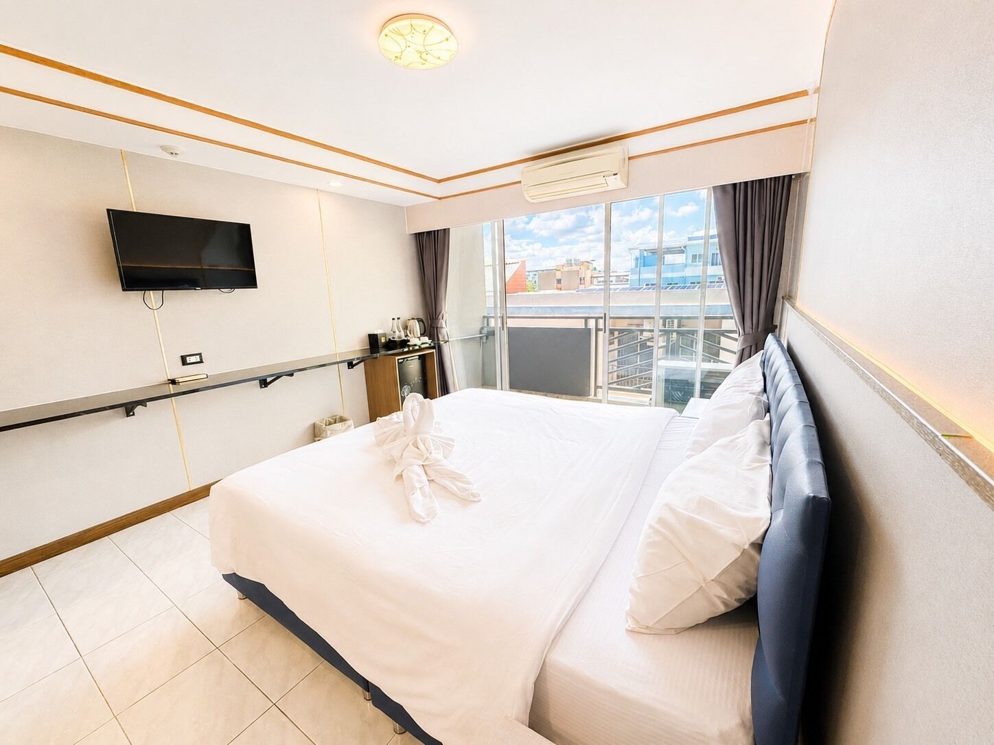 A hotel room with a bed, a TV mounted on the wall, a window with a view of buildings, and an air conditioning unit.