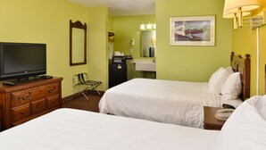 Ein Hotelzimmer mit zwei Betten, einem Fernseher, einem Schreibtisch mit Lampe, einem Spiegel und einem Bild an der Wand.