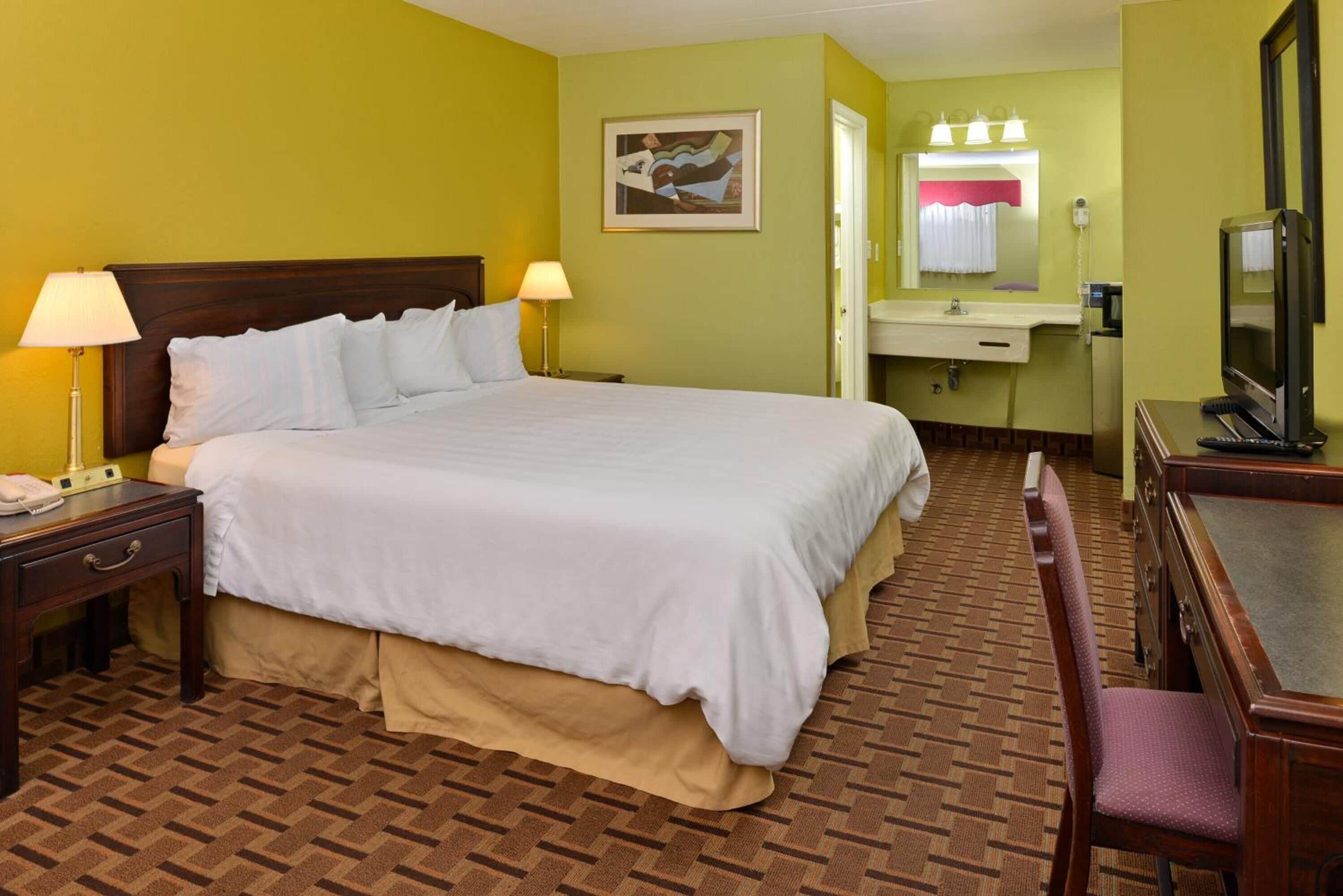 Habitación de hotel con una cama grande, lámparas de noche, un escritorio con teléfono, una silla, televisión y baño con lavabo y espejo.