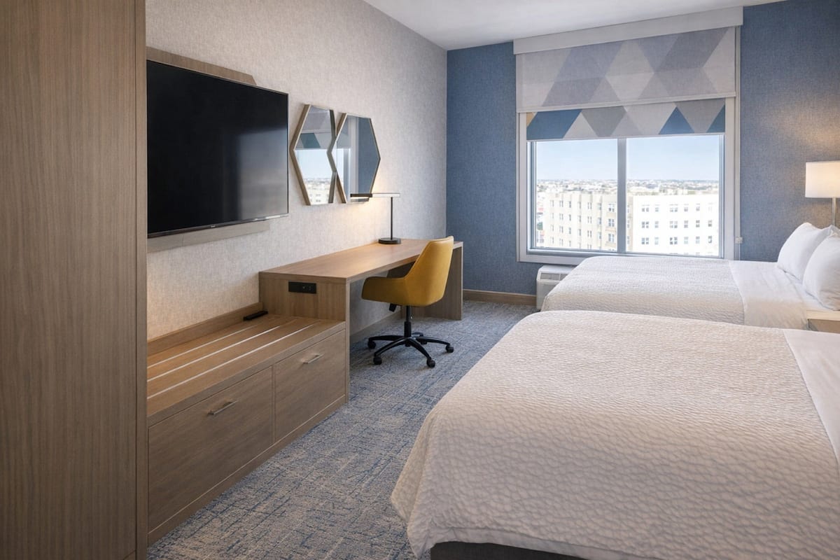 A hotel room with a bed, a desk with a chair, a TV, and a large window.