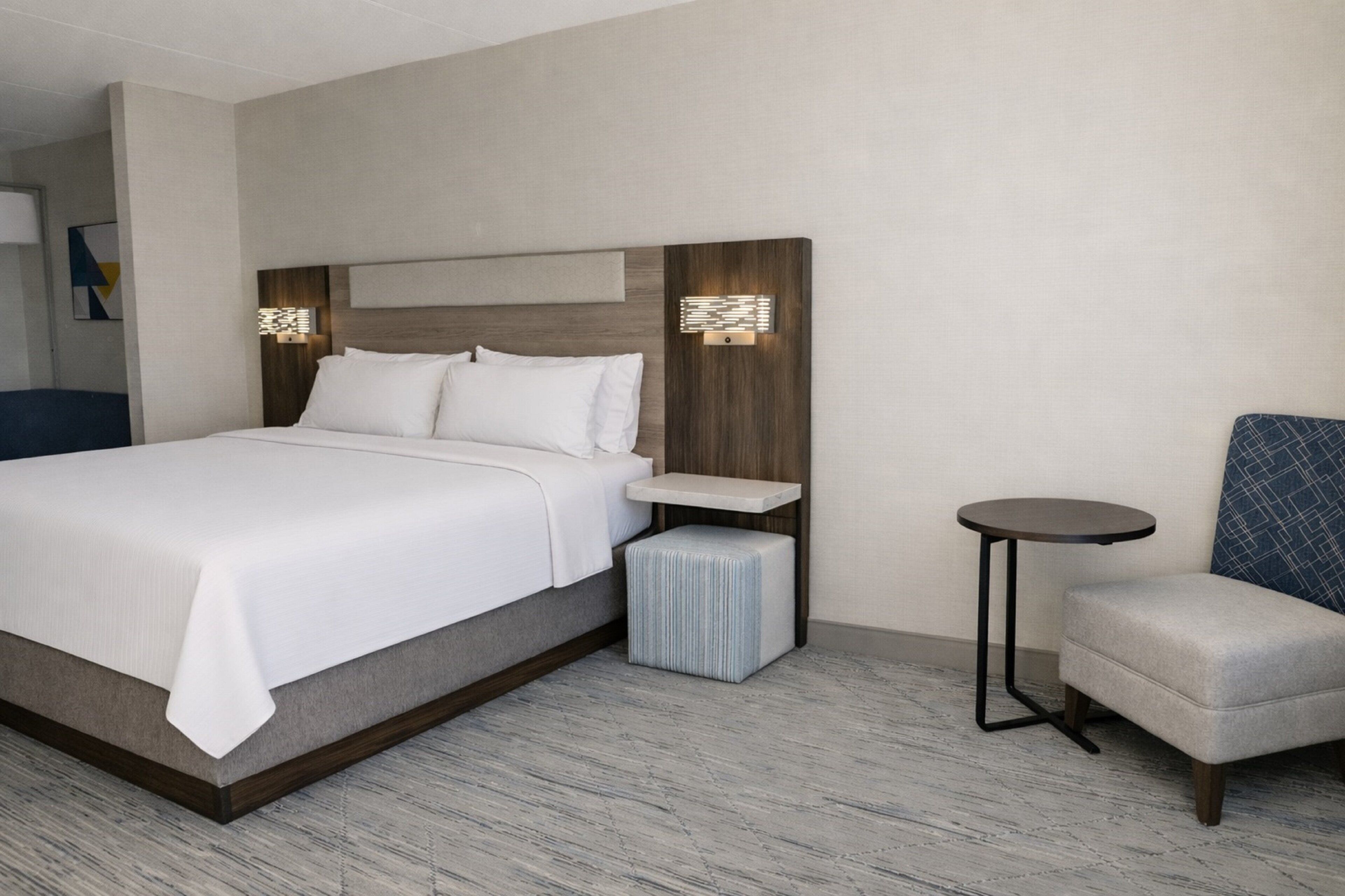 Una habitación de hotel con una cama grande, mesitas de noche, una silla y una pequeña mesa redonda.