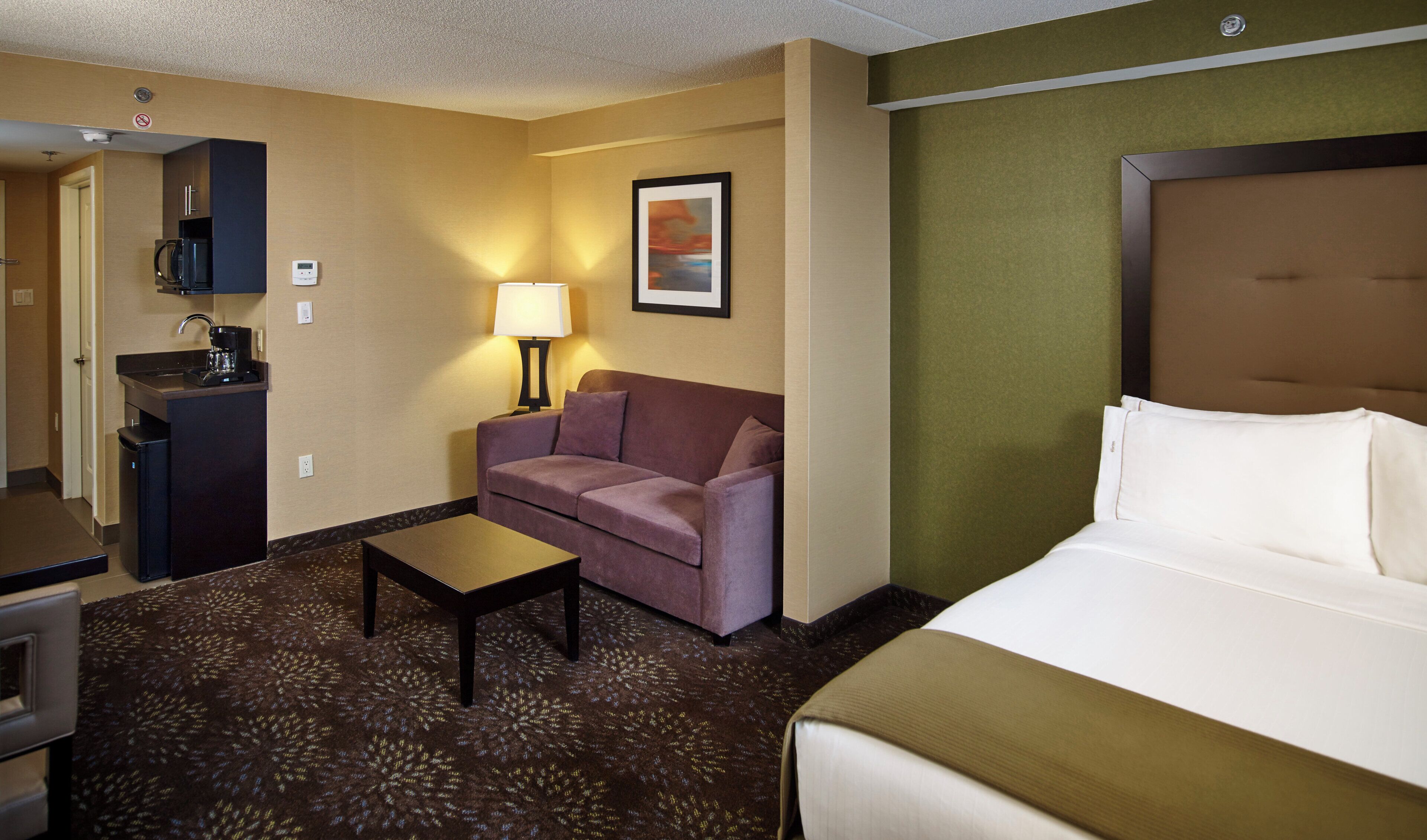A hotel room with a bed, a sofa, a coffee machine, a desk, and a lamp.