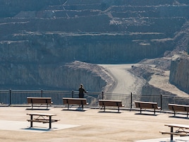 Una persona parada sobre una plataforma de concreto con bancos y mesas de picnic, con vista a un cañón profundo y un camino de tierra.