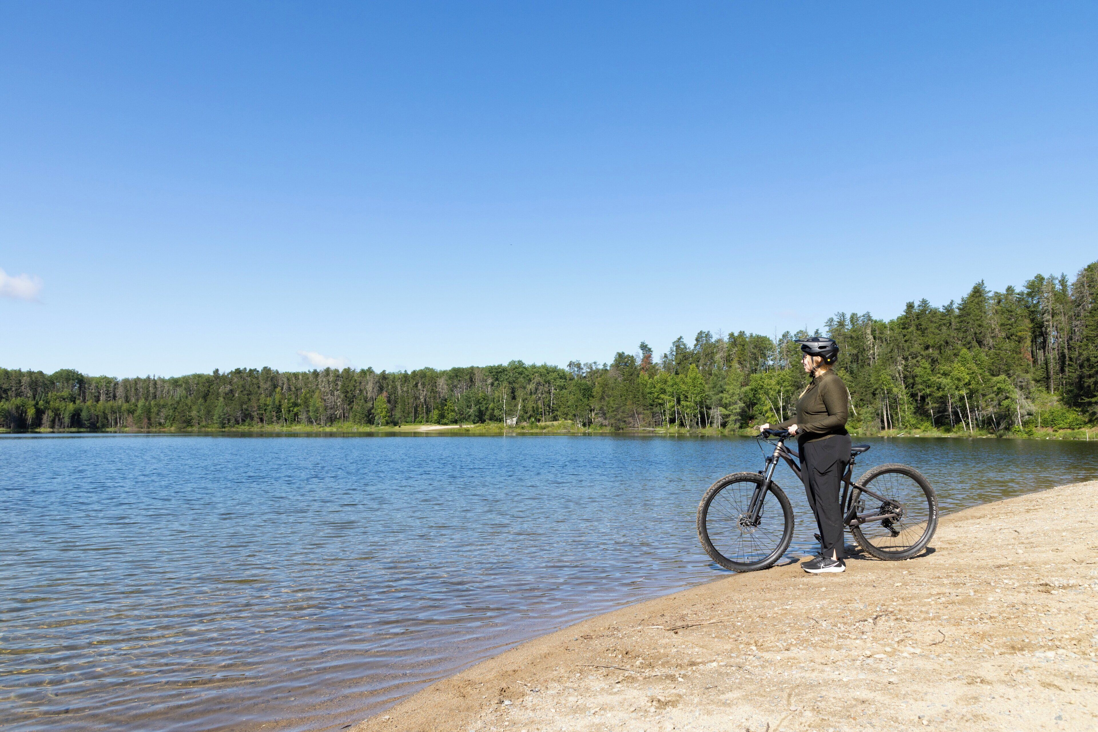 A person standing by a lake with a bicycle, surrounded by trees and clear skies.