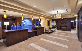 A hotel reception area with a reception desk, two receptionists, a wall-mounted TV, a fireplace, and a seating area with chairs and a table.