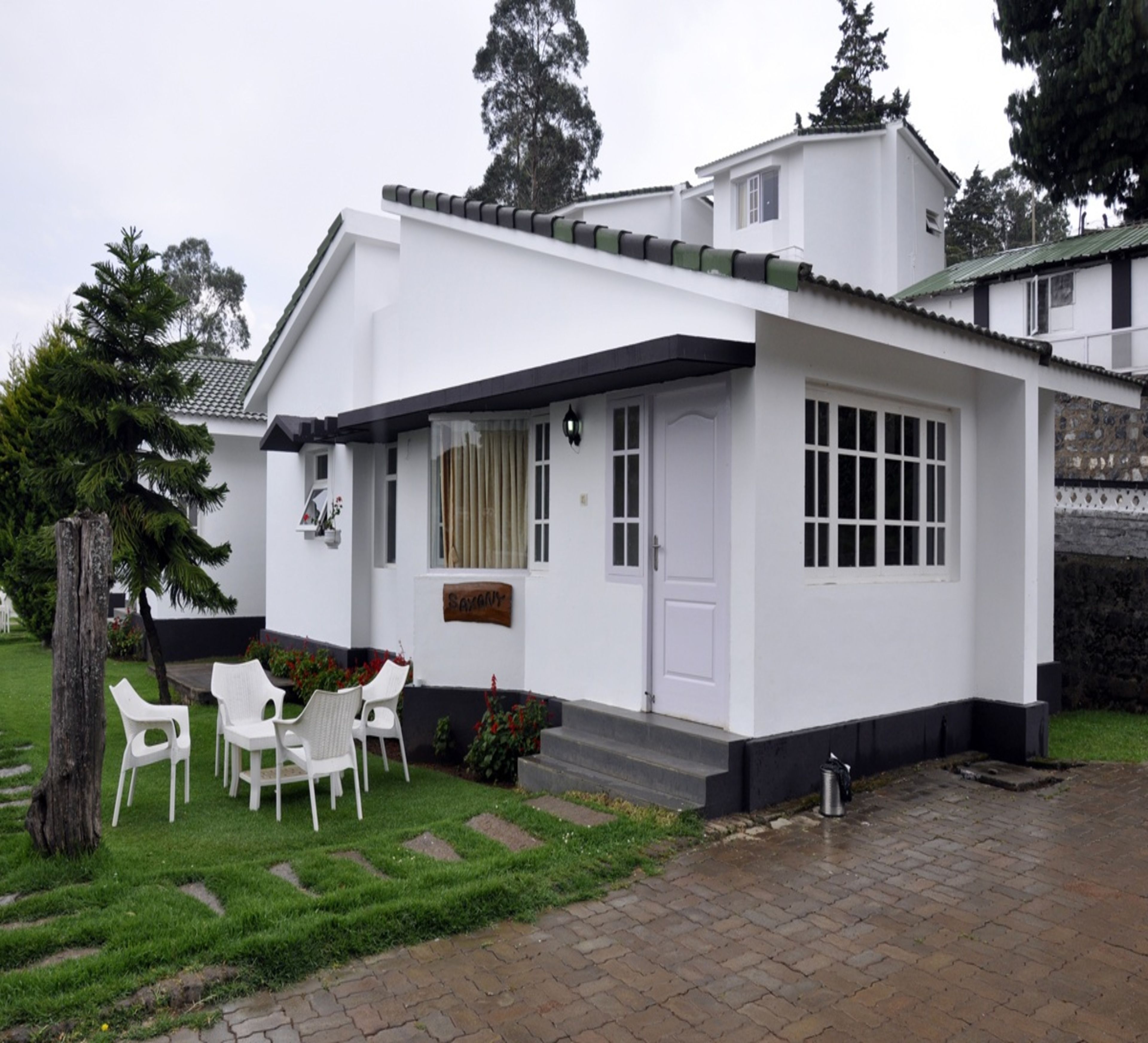 A white single-story house with a small porch, a door, and a window. There are white plastic chairs and a small table on the porch.