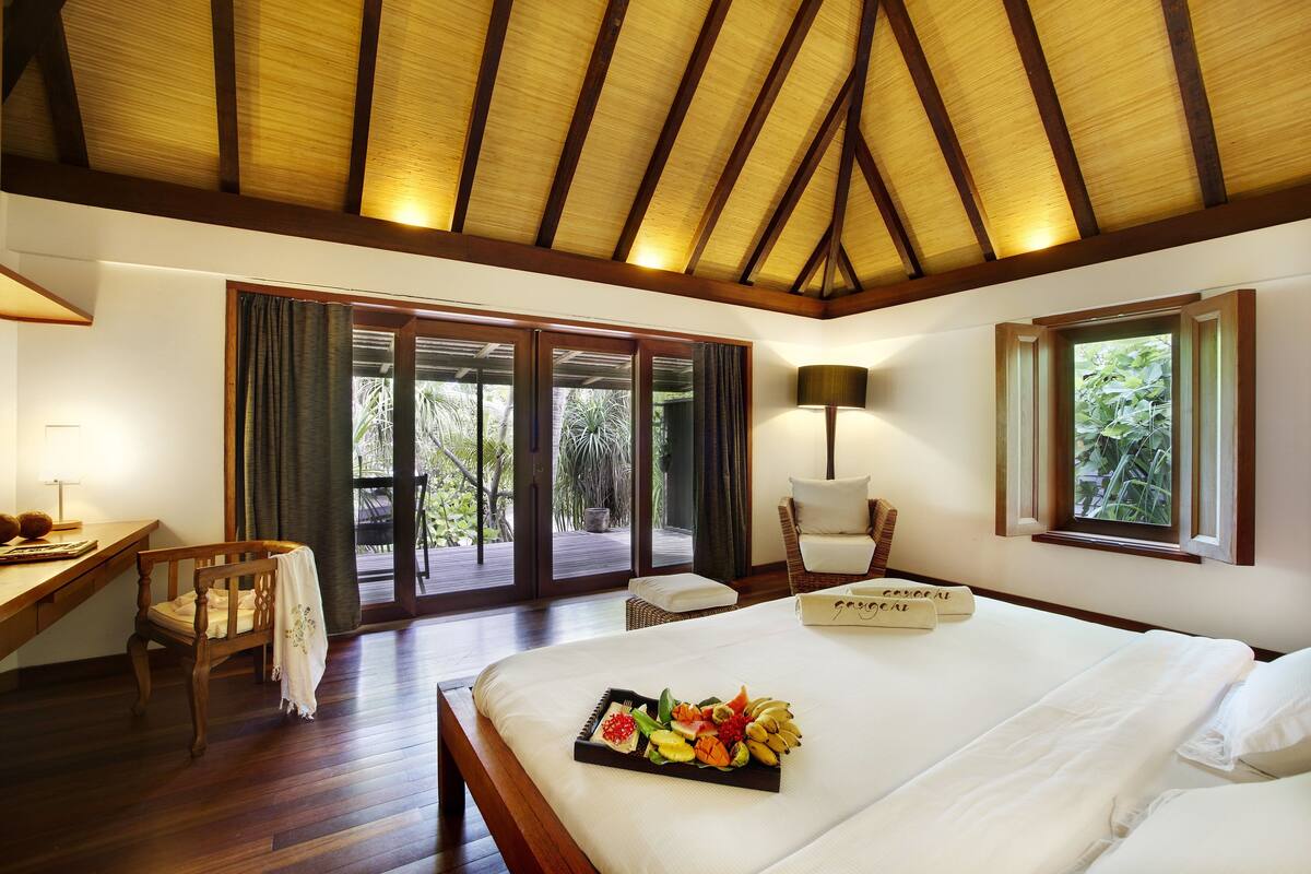 Habitación de hotel con techo de madera, una cama con una bandeja de fruta, un escritorio con lámpara y una silla.