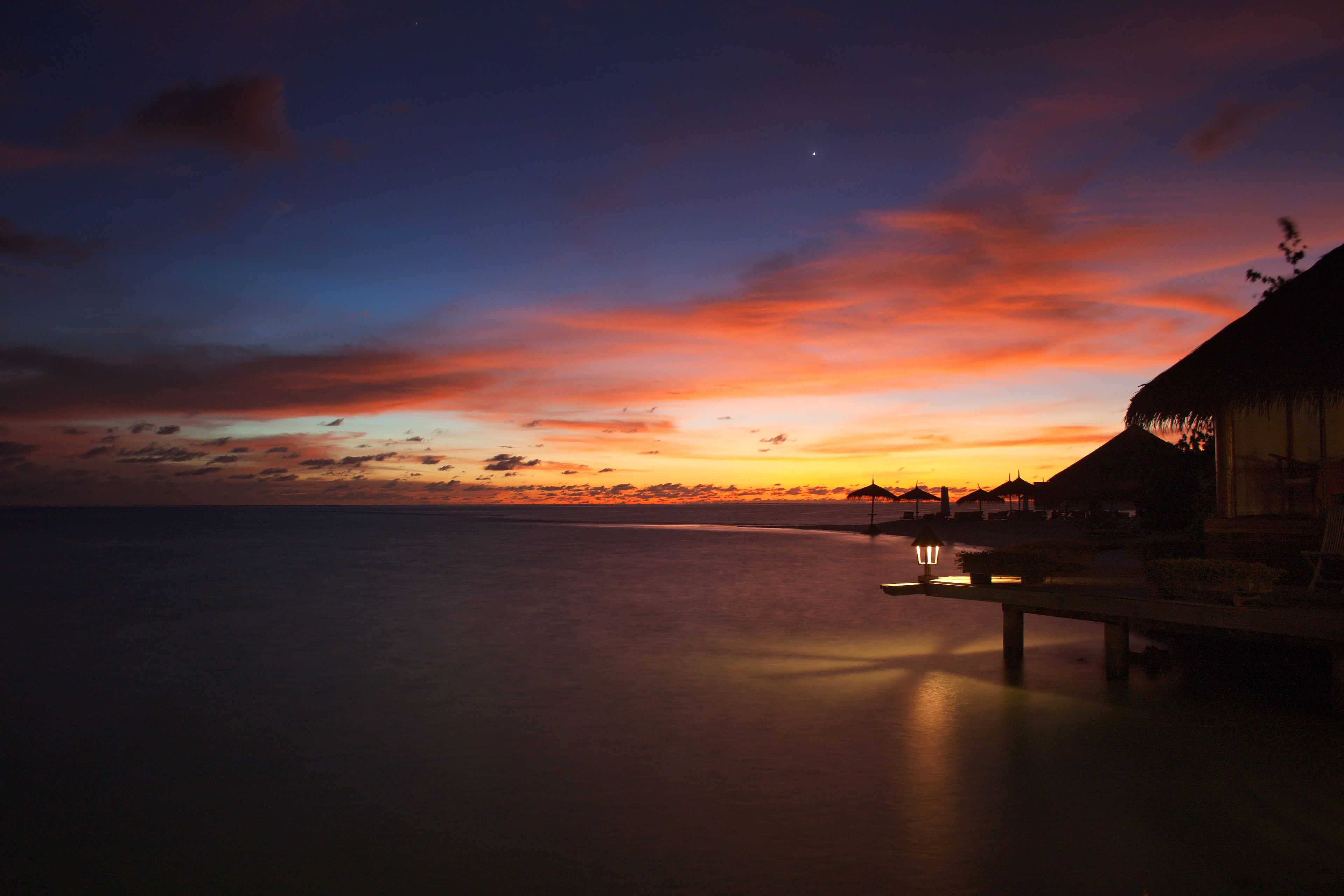 Un atardecer sobre un mar tranquilo con una estructura de techo de paja y un muelle que se adentra en el agua.