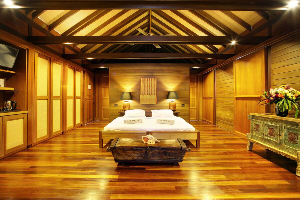 Una habitación de hotel con piso de madera, una cama con ropa blanca, cabecera de madera, mesita de noche con lámpara y un jarrón decorativo con flores.