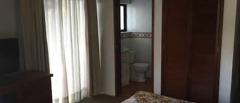 Habitación de hotel con cama, escritorio, espejo y baño visible a través de una puerta abierta.
