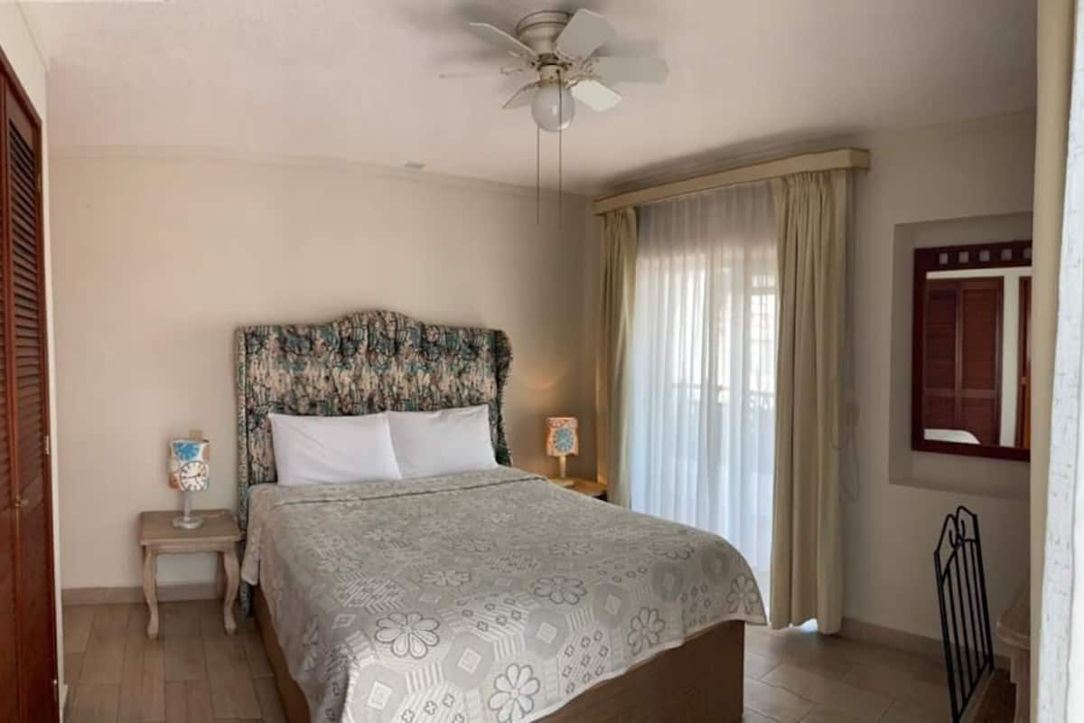 Een slaapkamer met een bed, een plafondventilator, een nachtkastje, een lamp, een raam met gordijnen en een deur.