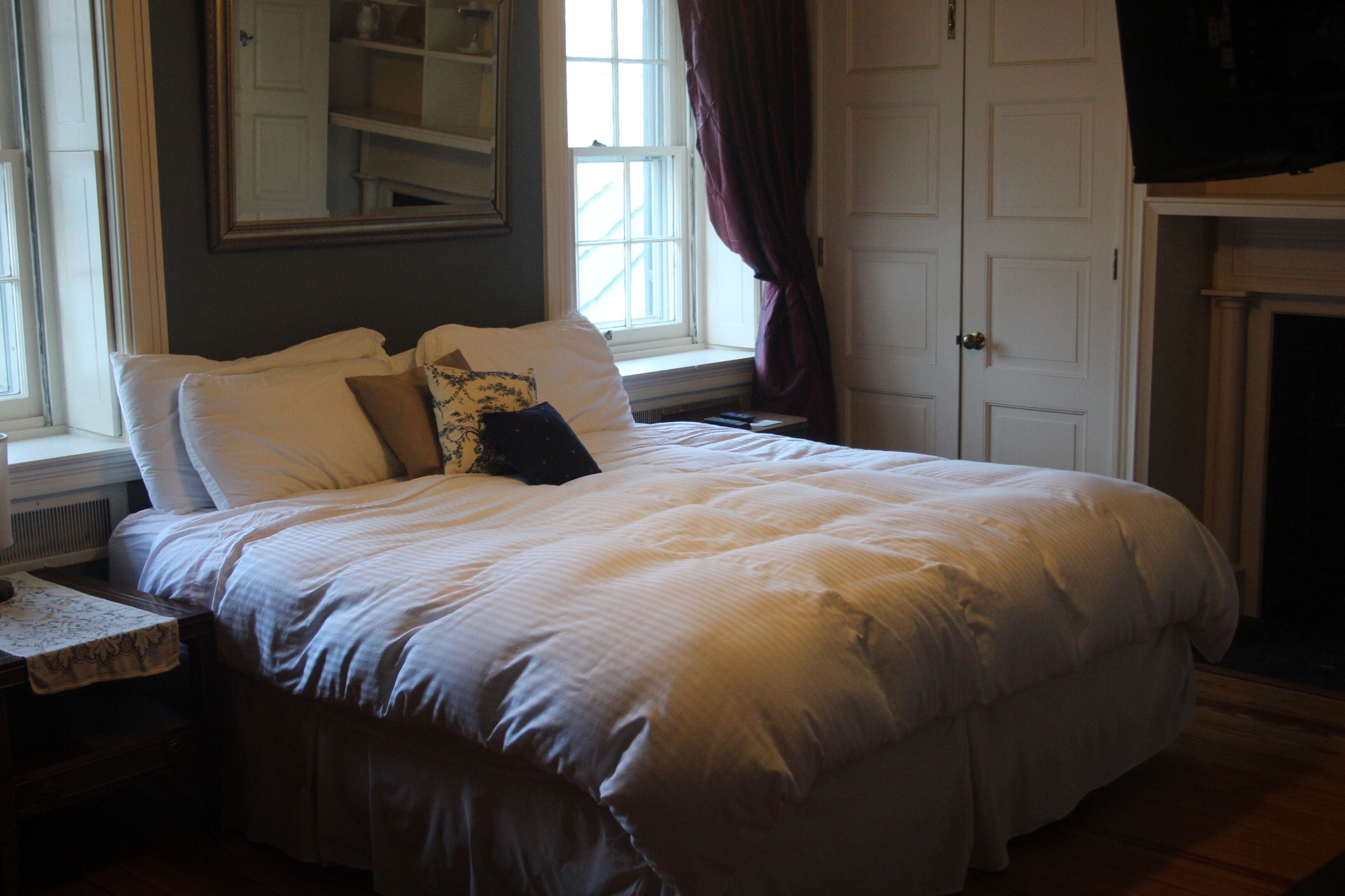 A neatly made bed with a white comforter and several pillows, a small bedside table with a decorative item, and a window with curtains.