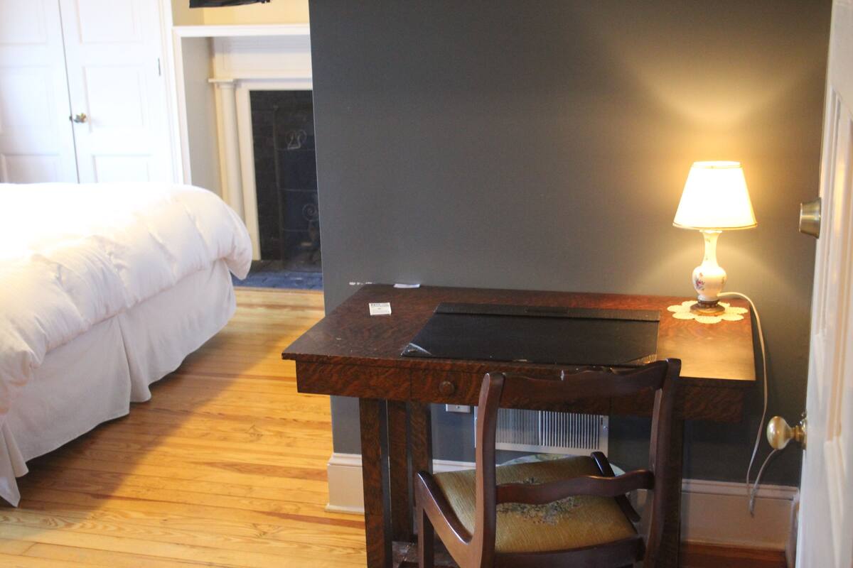 A hotel room with a wooden desk, a chair, a bed, and a lamp.