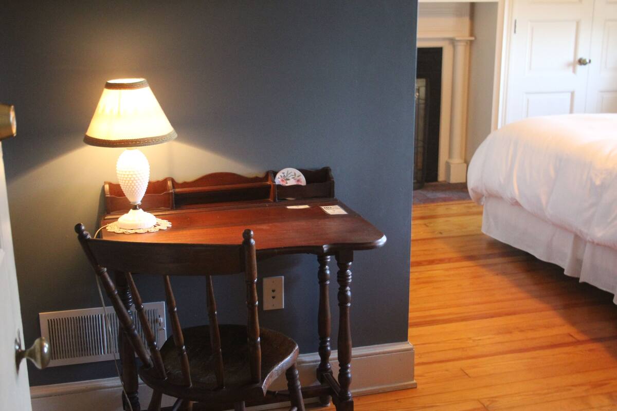 A room with a wooden table, a chair, a lamp, a bed, and a door.