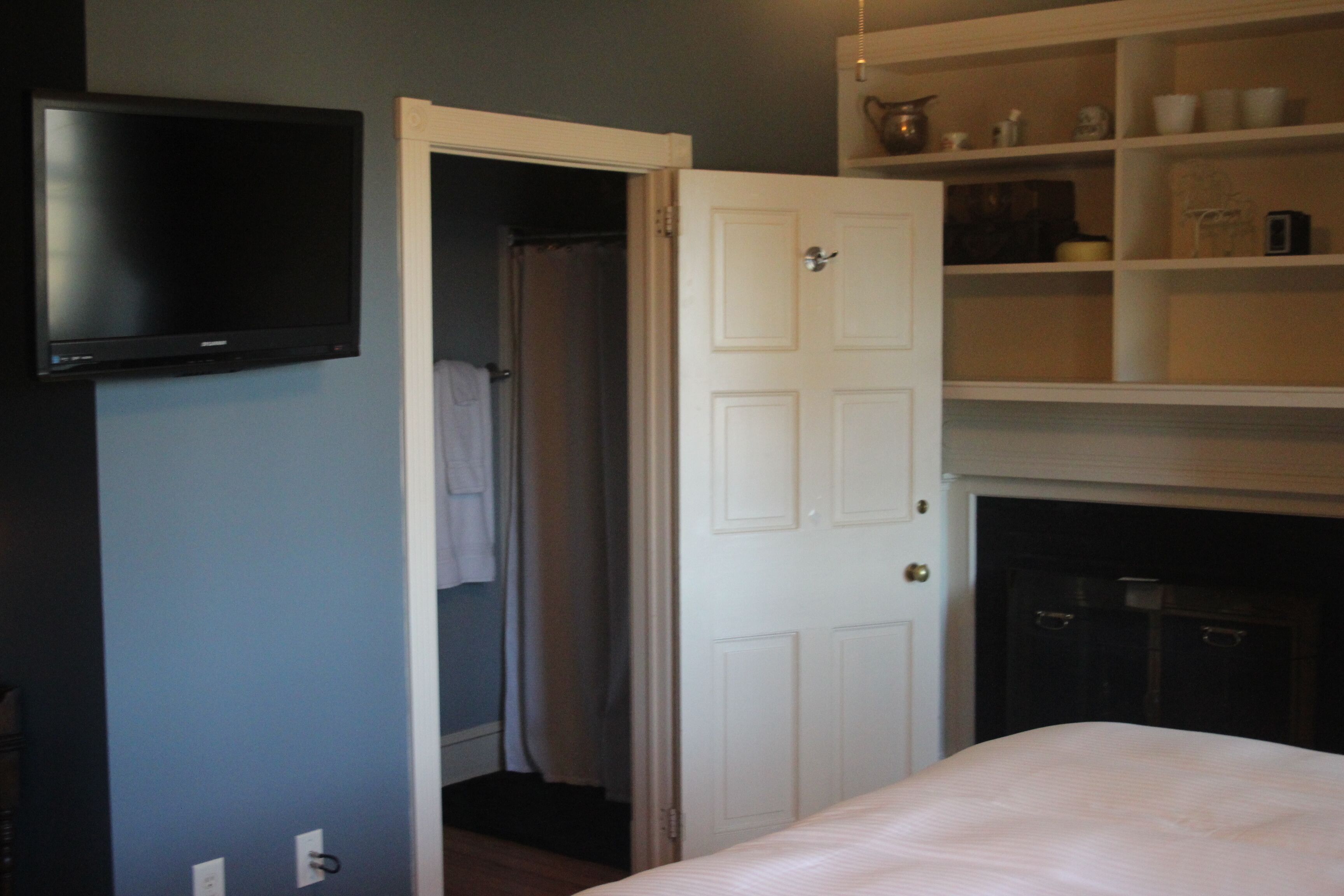 A bedroom with a bed, a television mounted on the wall, a white door, and a shelf with various items.