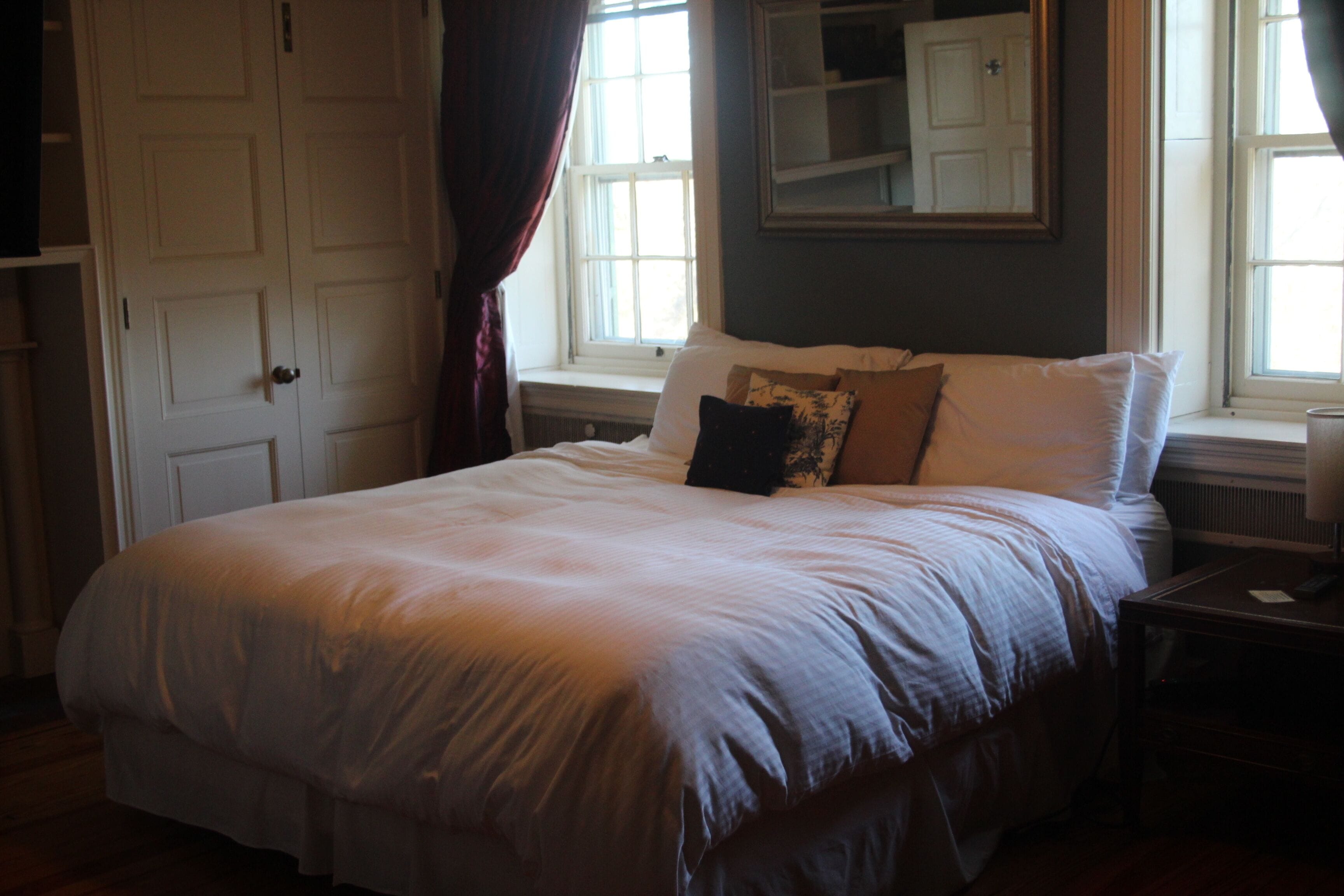 A neatly made bed with a white comforter and pillows, a bedside table with a remote control, and a window with curtains.