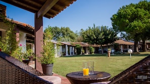 Un patio con mobiliario de mimbre, una mesa de vidrio con bebidas y vista a un patio interior con edificios y árboles.