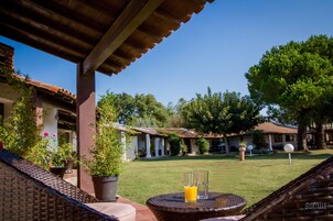 Un patio con mobiliario de mimbre, una mesa de vidrio con bebidas y vista a un patio interior con edificios y árboles.