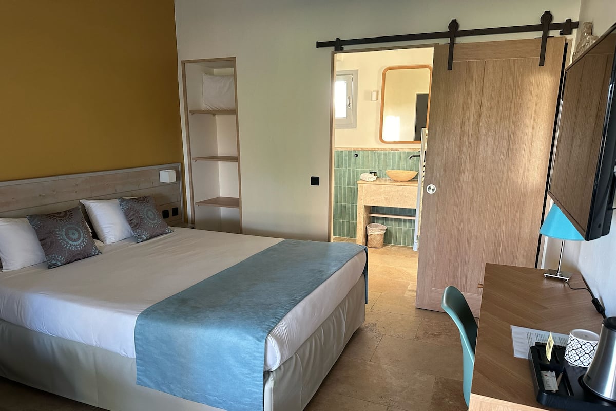 Ein Hotelzimmer mit einem großen Bett, einem Schreibtisch mit Lampe, einem Spiegel und einem durchgegangenen Türflügel sichtbar liegenden Badezimmer.