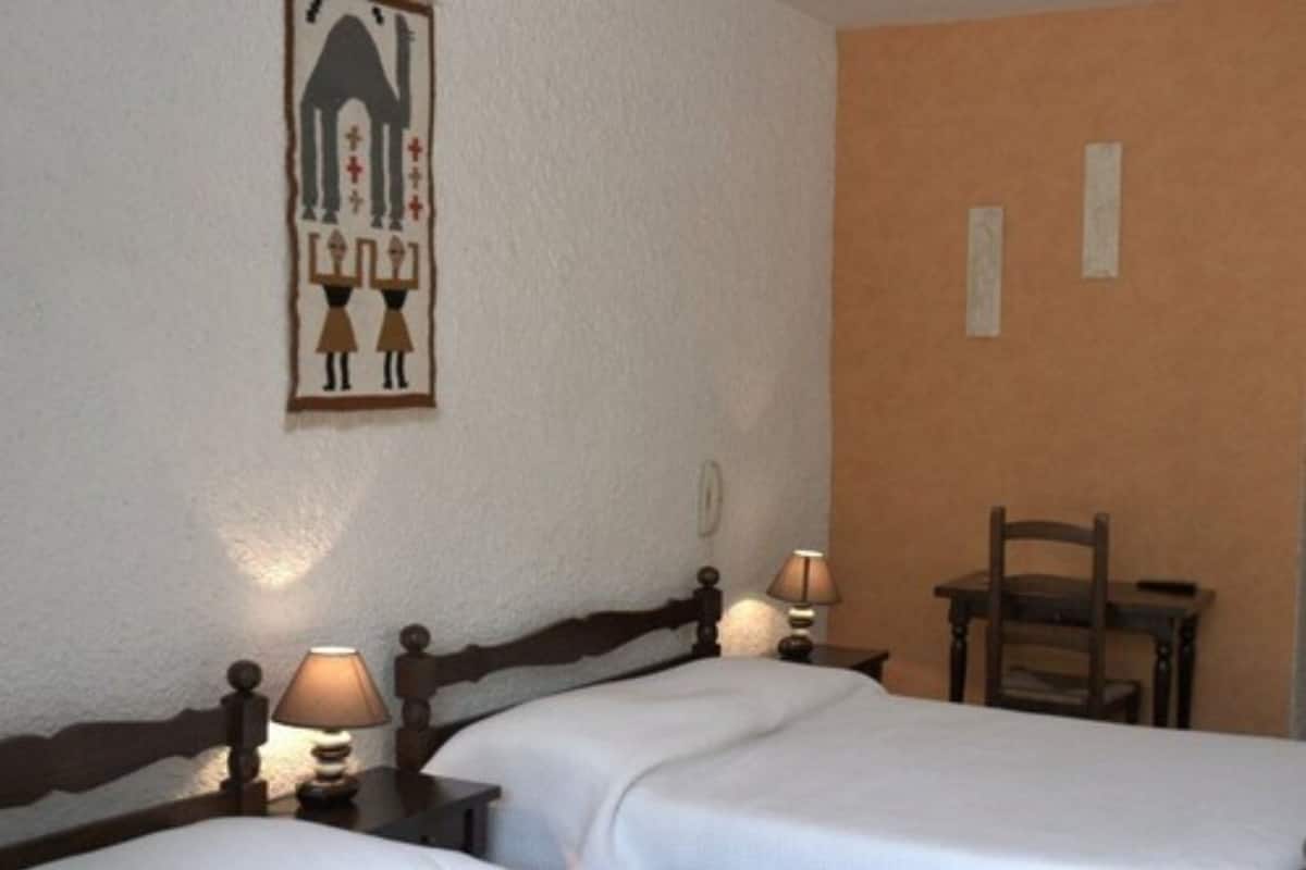 A hotel room with two beds, a wooden headboard, two bedside tables with lamps, a small wooden chair, and a wall-mounted picture.