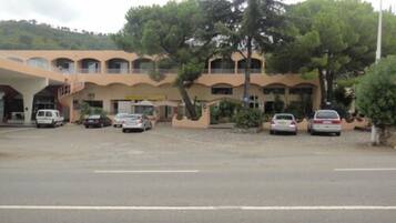 A multi-story hotel with a central courtyard, several cars parked in front, and a large tree providing shade.