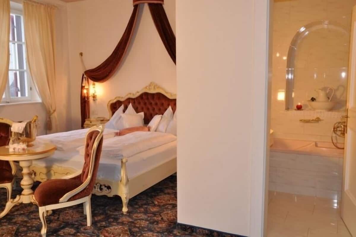 Une chambre dotée d’un grand lit, d’un coin salon avec une table et une chaise, et d’une salle de bain avec une baignoire et un miroir.