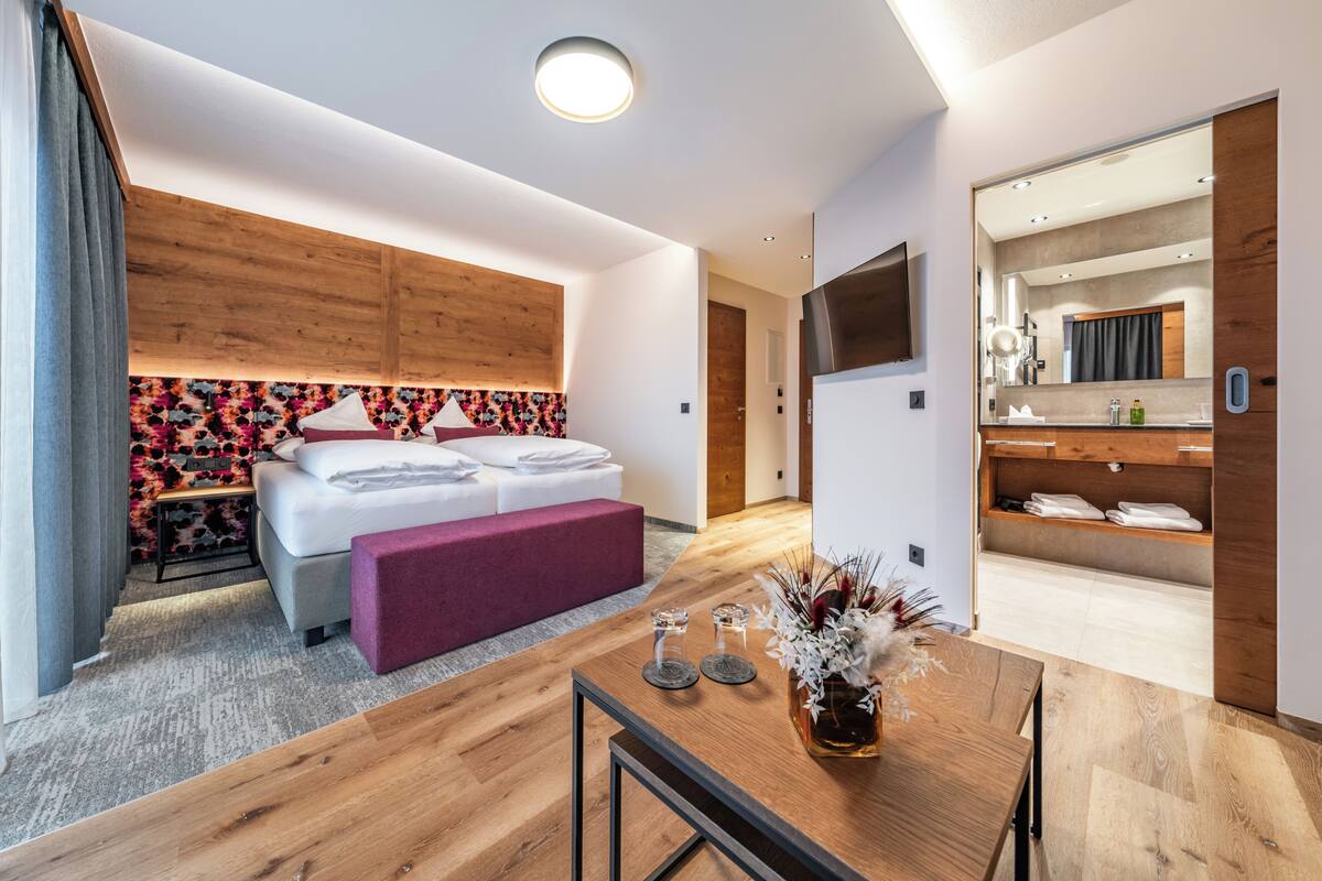 A modern hotel room with a wooden headboard, a bed with white linens, a purple bench, a wooden dining table with a vase, and a bathroom with a mirror and sink.