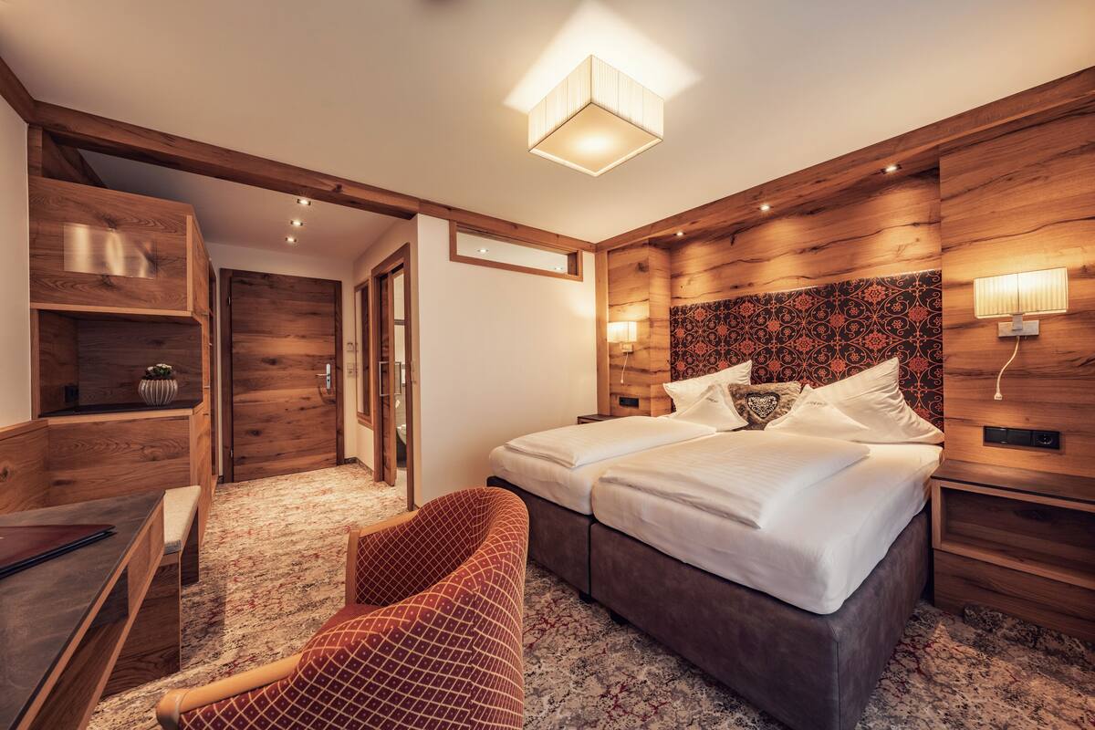 A hotel room with a wooden headboard, two beds, a bench, a desk, and a patterned carpet.