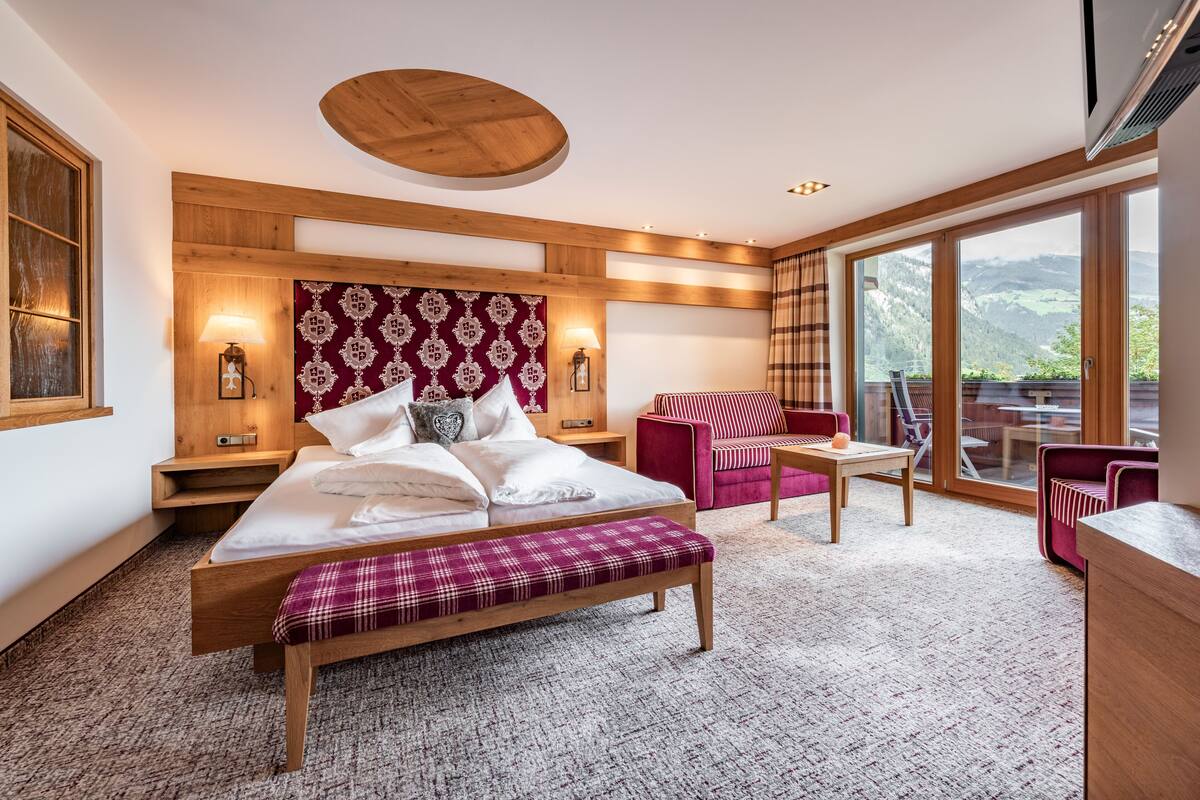 A modern hotel room with a wooden bed, a red and white patterned headboard, a large window with a mountain view, and a balcony with red chairs.