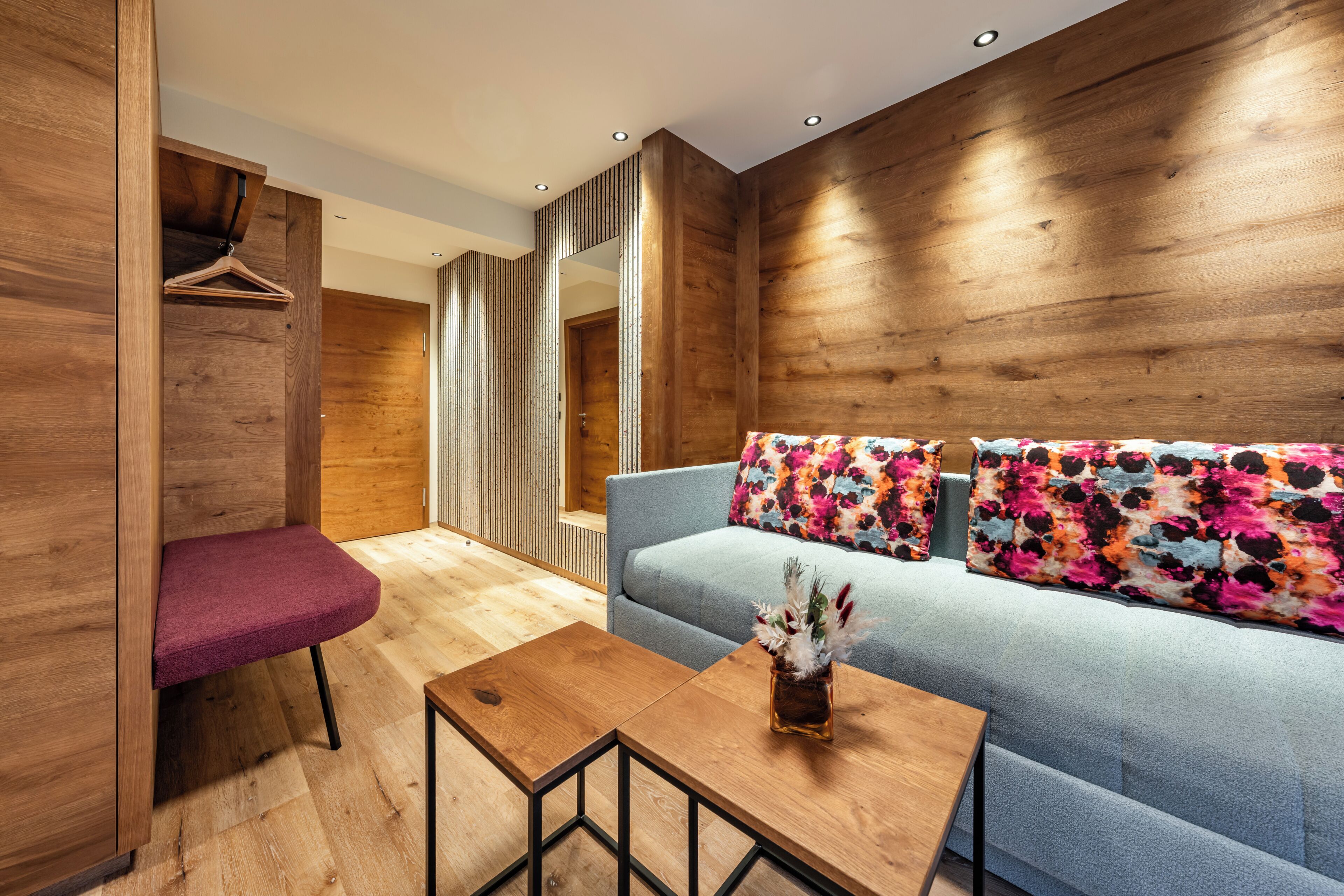 A modern room with a wooden wall, a blue sofa with colorful pillows, a wooden table, and a hanging rack.