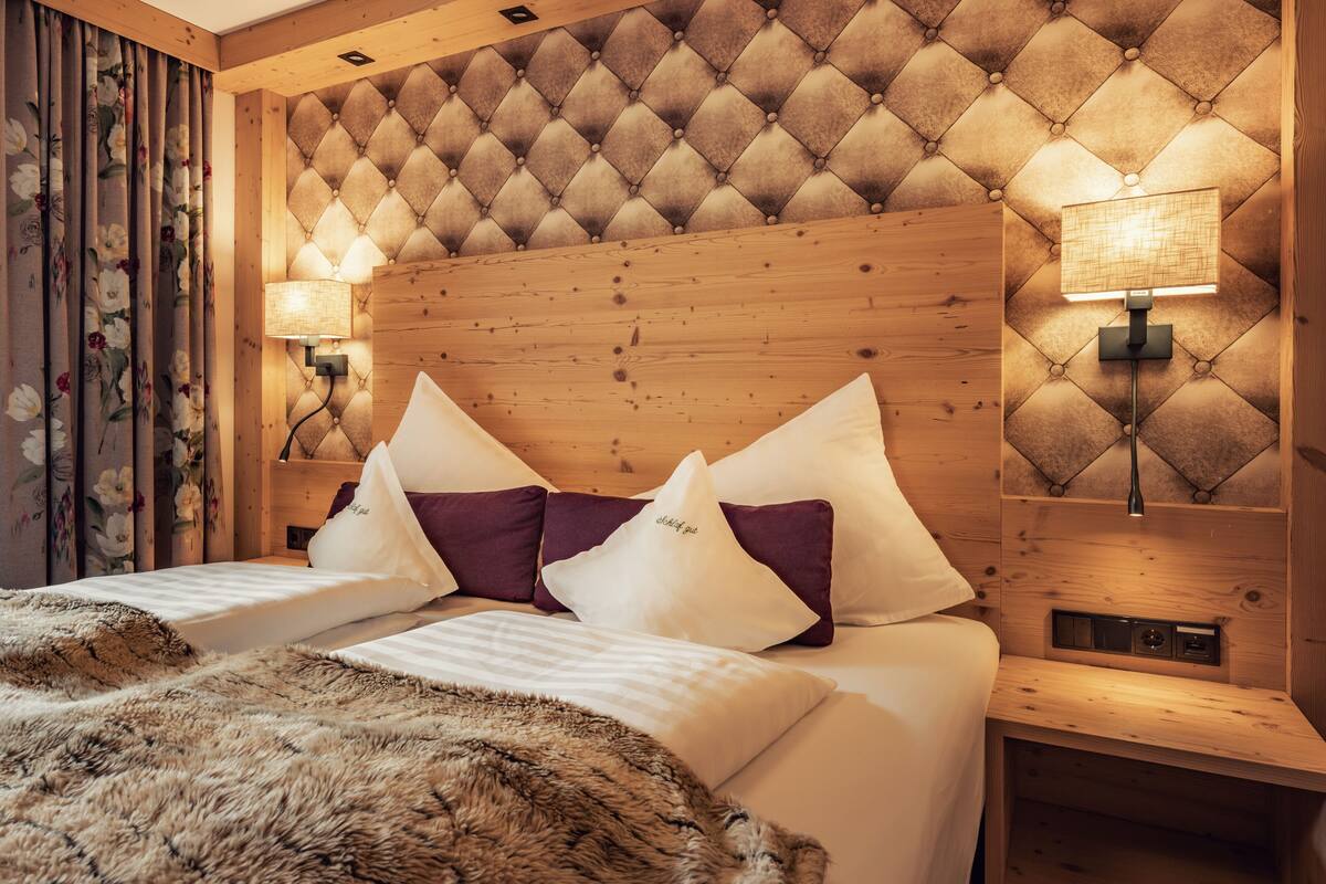 A hotel room with a tufted headboard, two bedside tables with lamps, a bed with white linens and purple pillows, and a wooden nightstand.