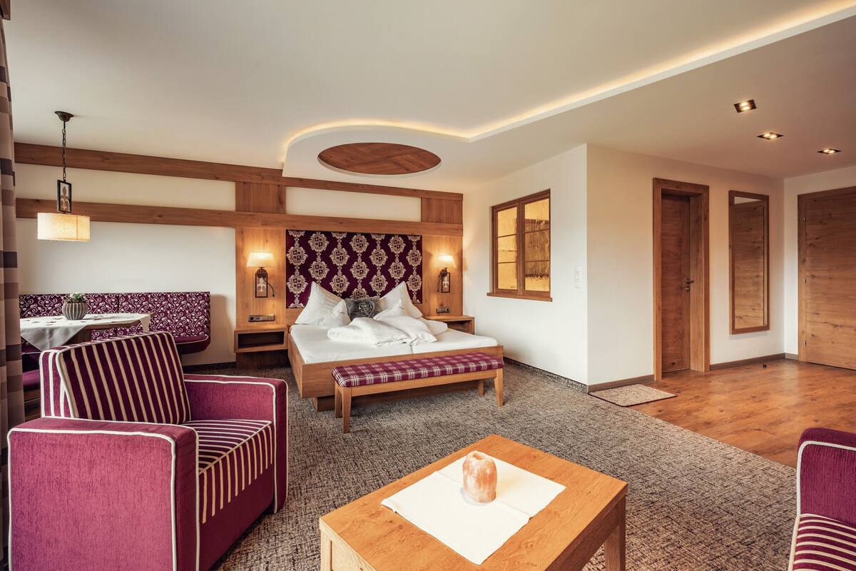A modern hotel room with a wooden ceiling, a large bed with a patterned headboard, a seating area with striped chairs, and a small table with a salt lamp.