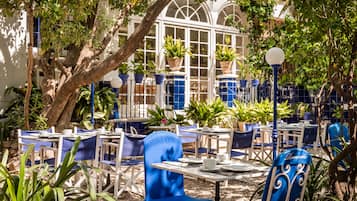 An outdoor café with tables and chairs, surrounded by potted plants and trees.