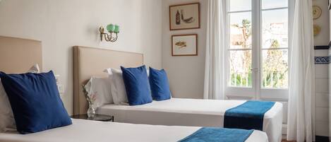 A hotel room with two beds, each with a white and blue bedspread, a small table with a vase, and a window with curtains.