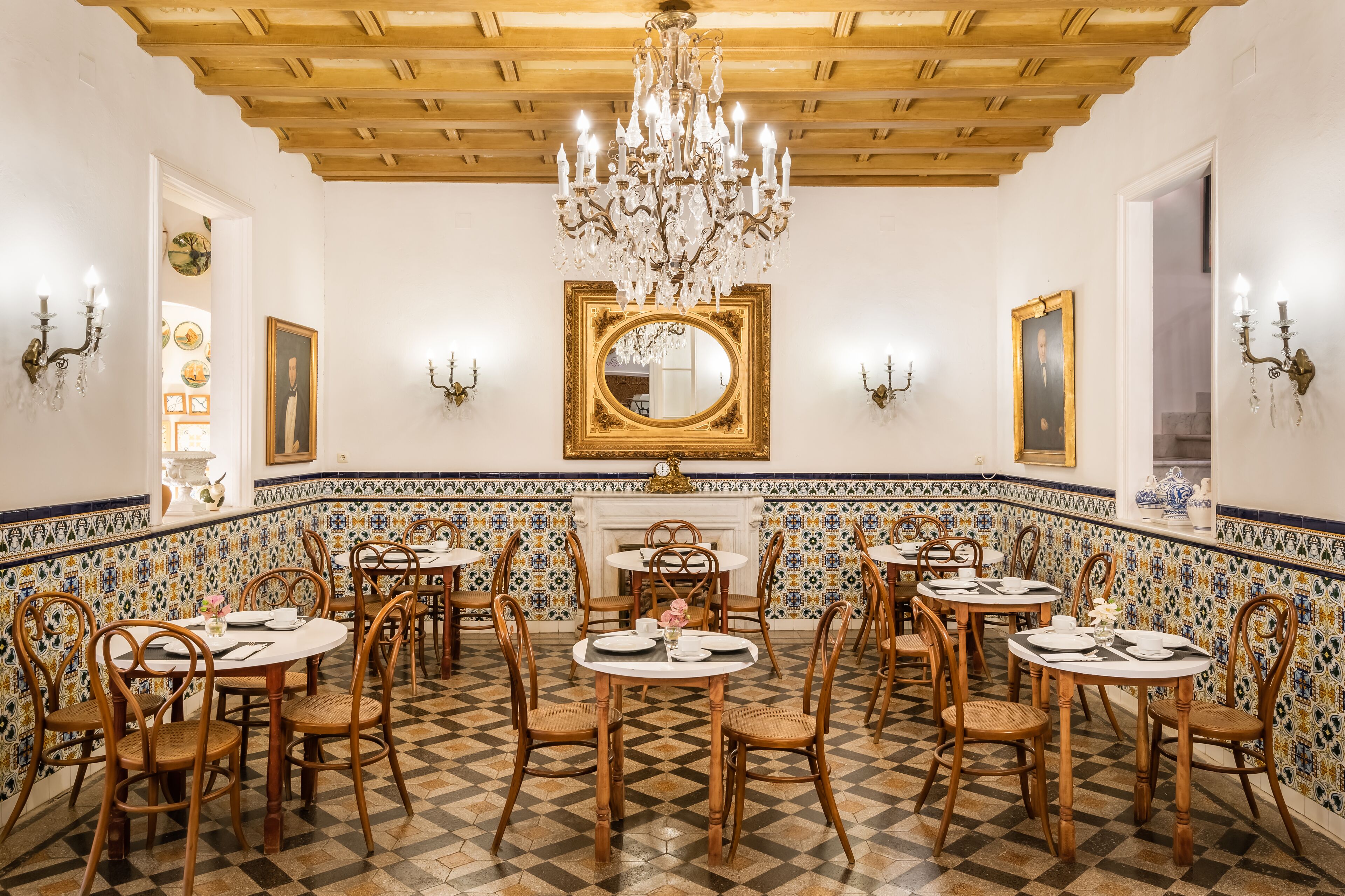A dining area with round tables, wooden chairs, a chandelier, and a tiled floor.