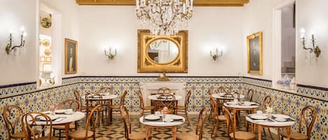A dining area with round tables, wooden chairs, a chandelier, and a tiled floor.